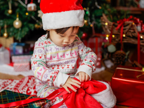 toddler baby girl wearing santa claus costume looking for gift in front of christmas tree - Image ; Mcimage /Shutterstock