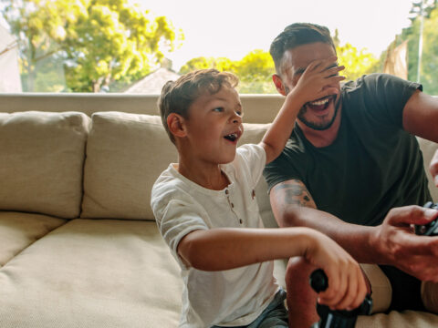Little boy covering eyes of his father playing video game. Cheerful family of father and son having fun playing video games at home. Courtesy Jacob Lund/Shutterstock