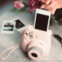 Pink instant camera printing a blank photo on a table with other photos and pink roses