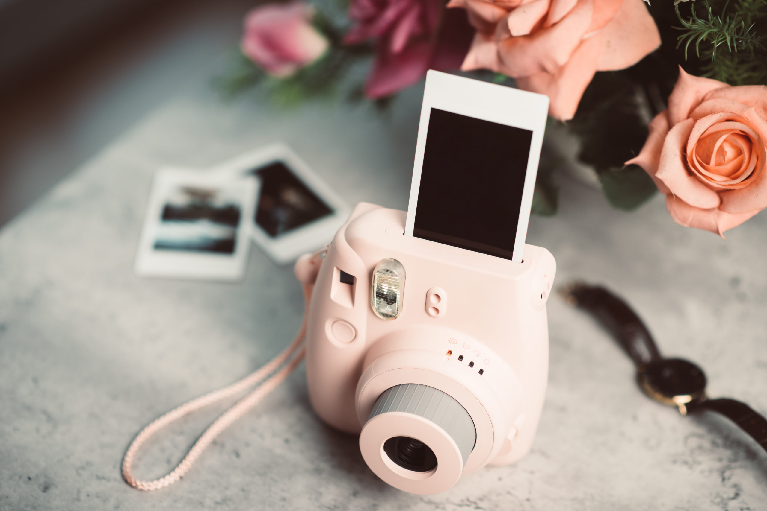 Pink instant camera printing a blank photo on a table with other photos and pink roses