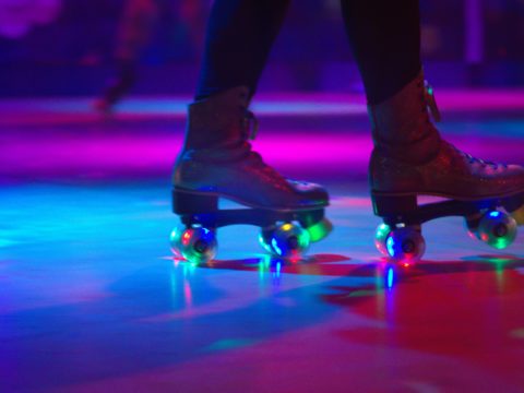A person from the shins down, wearing a pair of roller skates in a dark roller rink with pink and purple lights