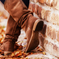 Person wearing tall fall boots, leaning against a brick wall and standing on fallen leaves